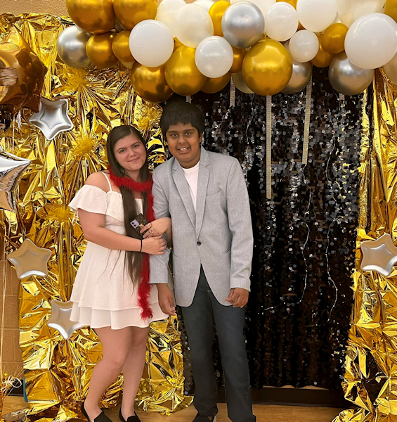 Photo of a girl and boy in dressy clothing standing underneath a balloon arch at a reception before a school dance.