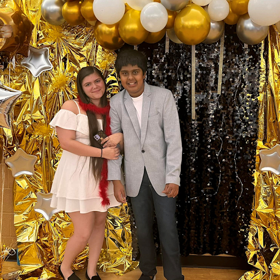 Two young teenagers dressed in fancy clothes, their arms interlocked, pose together at a party against a backdrop of black, gold, and silver sparkles and stars.  Above them is a balloon arch.