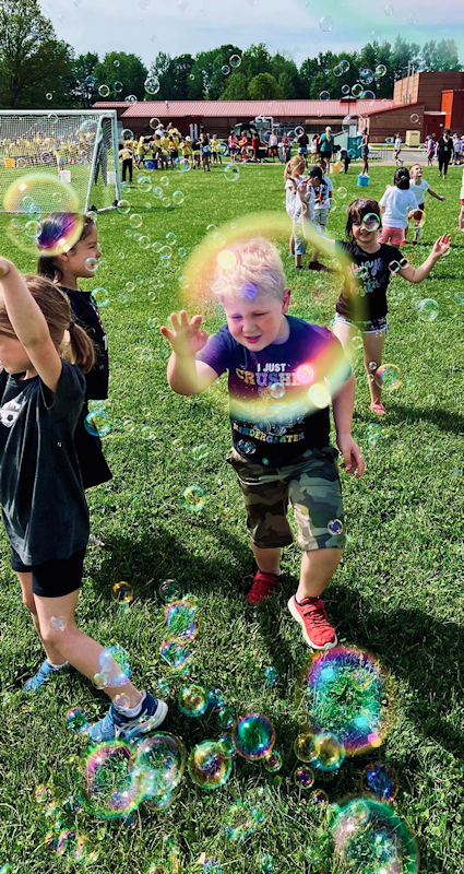 Photo of kindergarten children chasing bubbles in a field outside the school. One boy, who is wearing a shirt that says I crushed kindergarten, is seen through a large bubble.