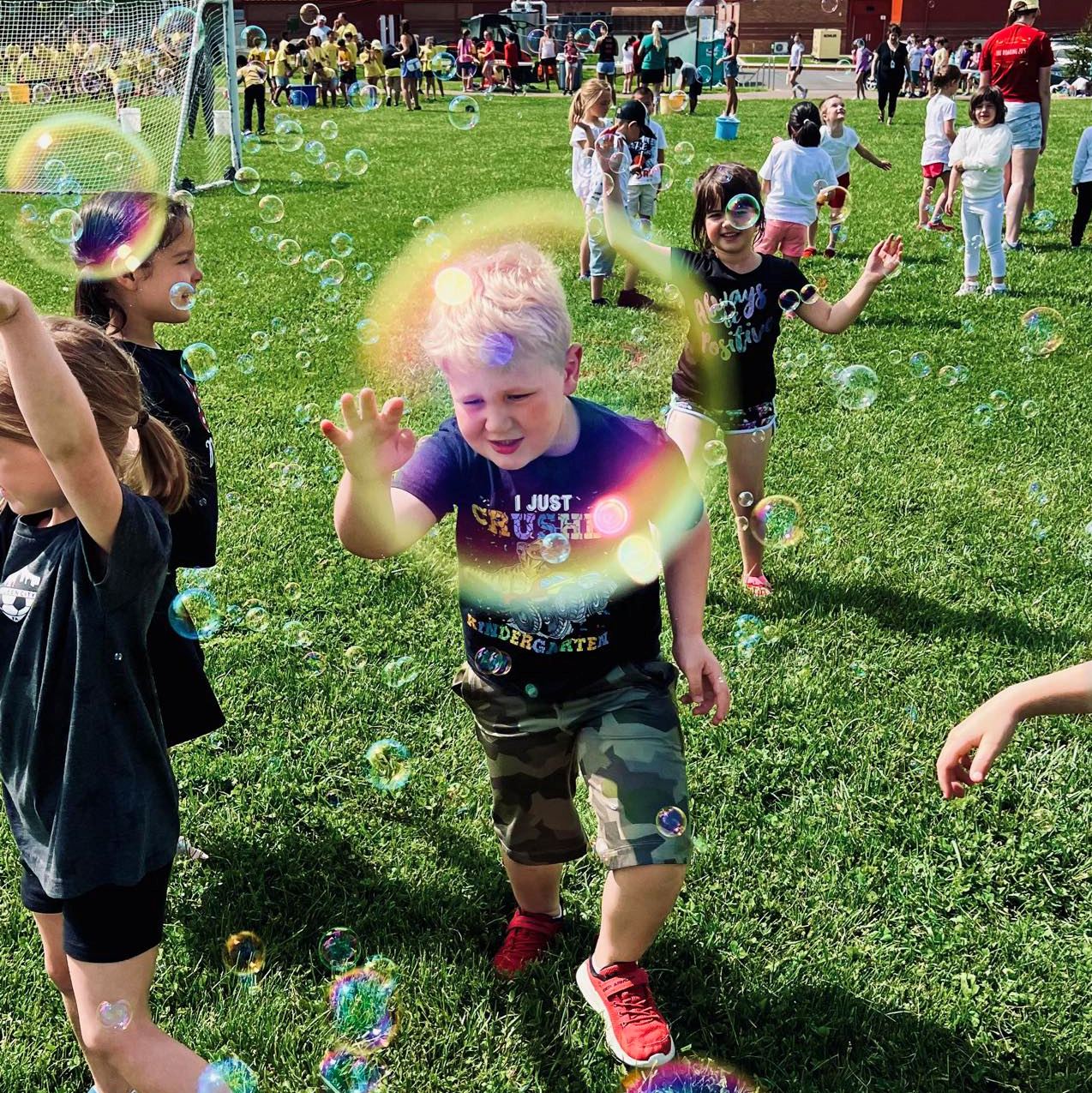 Groups of children are playing outdoors in a school field.  Some are chasing floating bubbles. In the foreground, a boy's face is framed inside a bubble.  He wears a shirt saying 'I just crushed kindergarten.' Just behind him is a smiling girl wearing a shirt that reads 'Always be positive.'
