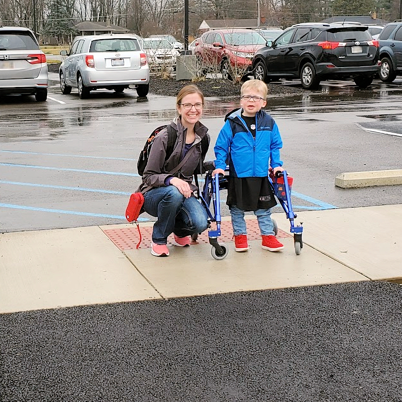 A smiling mom kneels next to her young son on a sidewalk square in a school parking lot.  The boy, who is wearing bright red shoes and a large sports jersey under a blue jacket, supports himself with a posterior pediatric walker. There is a blue striped vehicle access area behind them and a newly-completed asphalt ramp down from the sidewalk in front of them.